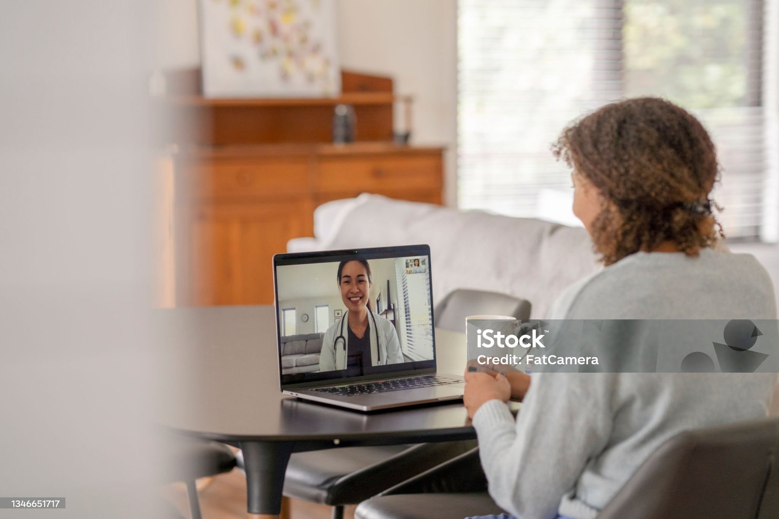 A female patent of African decent meets with her doctor remotely via a video call.  She is sitting at her kitchen table with her laptop out and a cup of coffee on the table with her.  The doctor can be seen on the screen wearing a white lab coat as she talks with her patient.