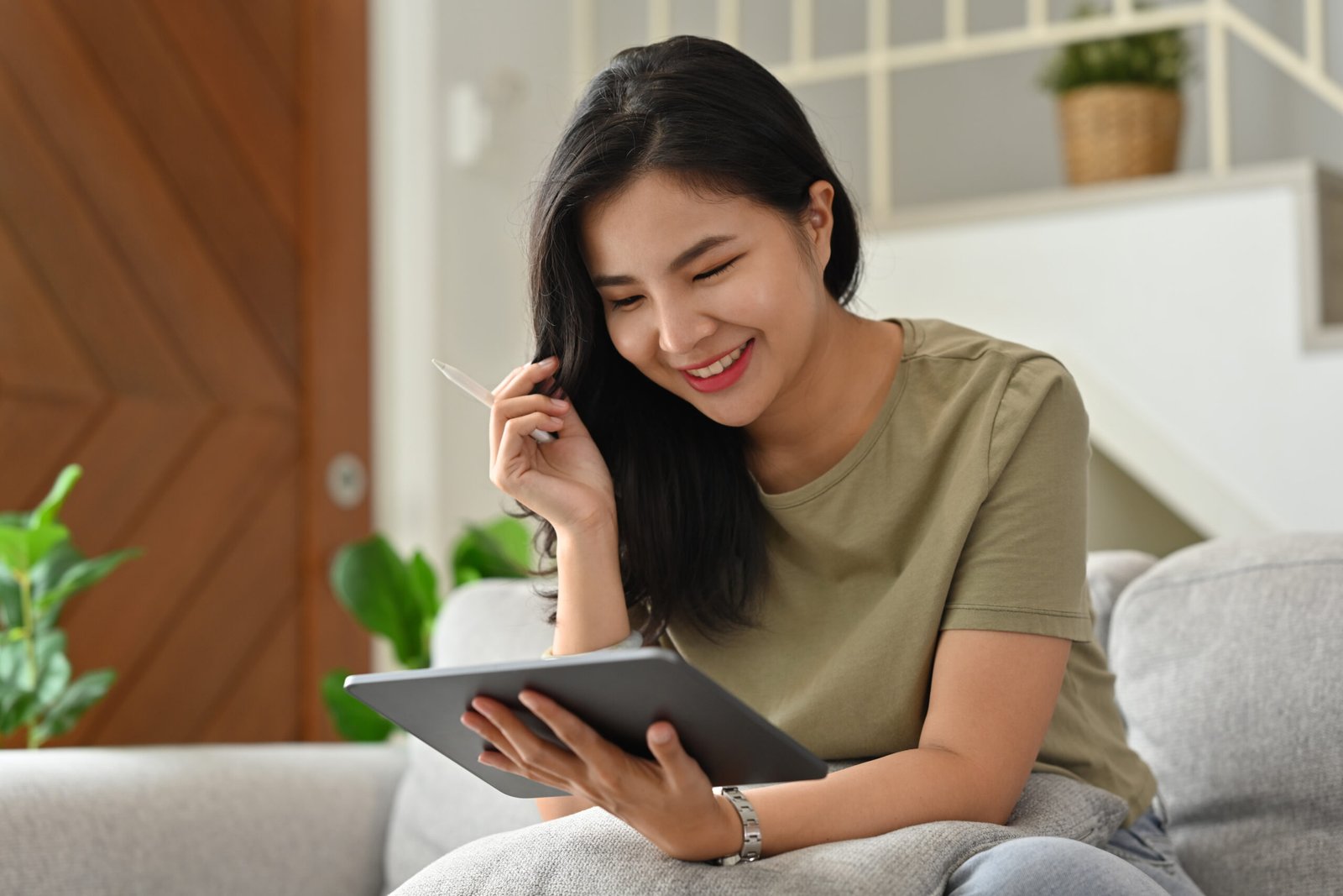 Domestic life and Sweet home concept with Millennial Asian woman using digital tablet and stylus pen while sitting on sofa.