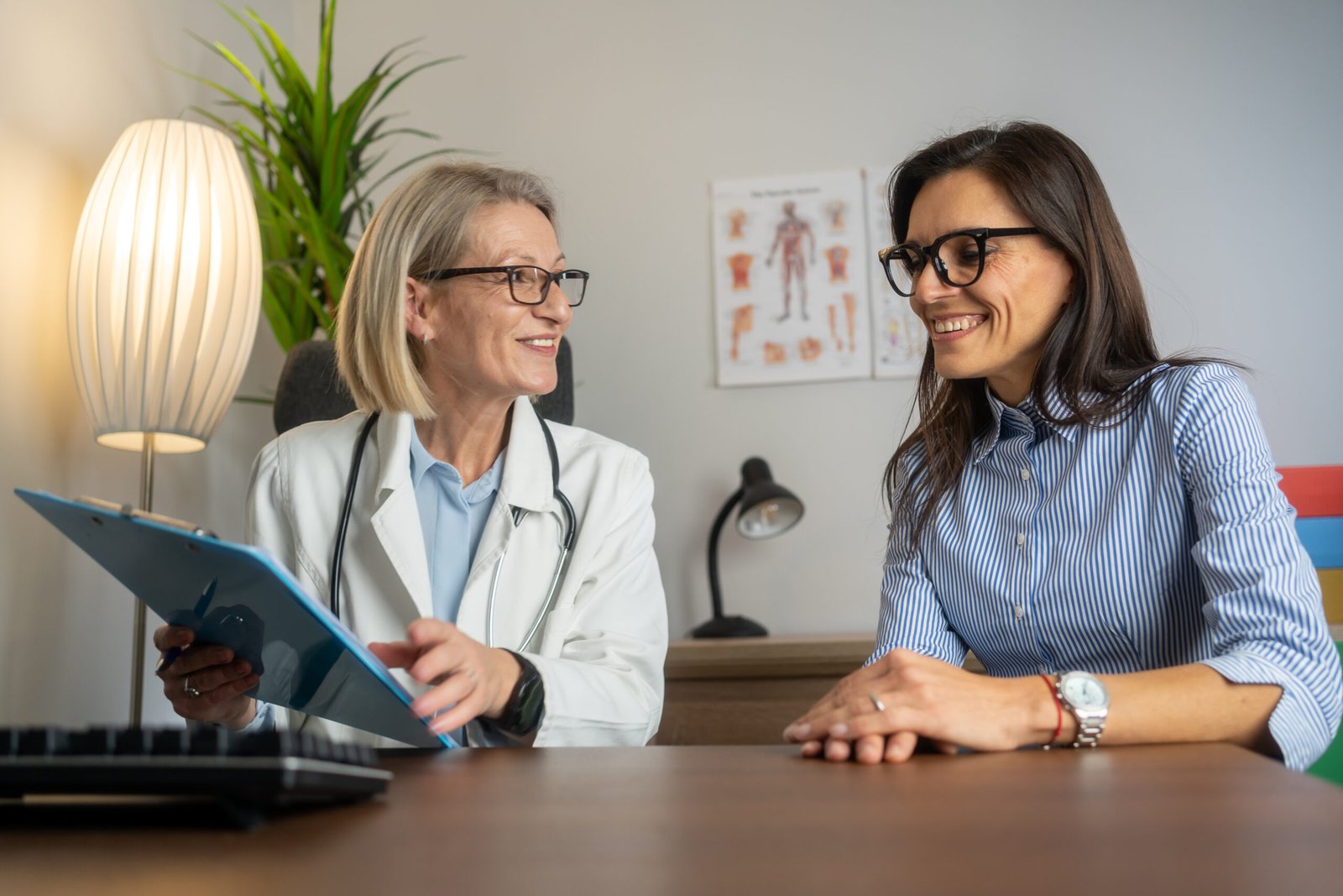 Smiling female doctor with clipboard talking with middle aged female patient in doctor office. Doctor questioning patient and writing answers in clipboard. medicine, healthcare and people concept