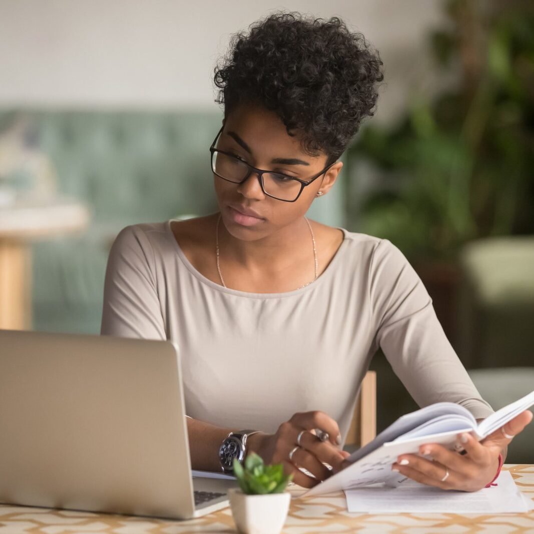 Focused young african american businesswoman or student looking at laptop holding book learning, serious black woman working or studying with computer doing research or preparing for exam online